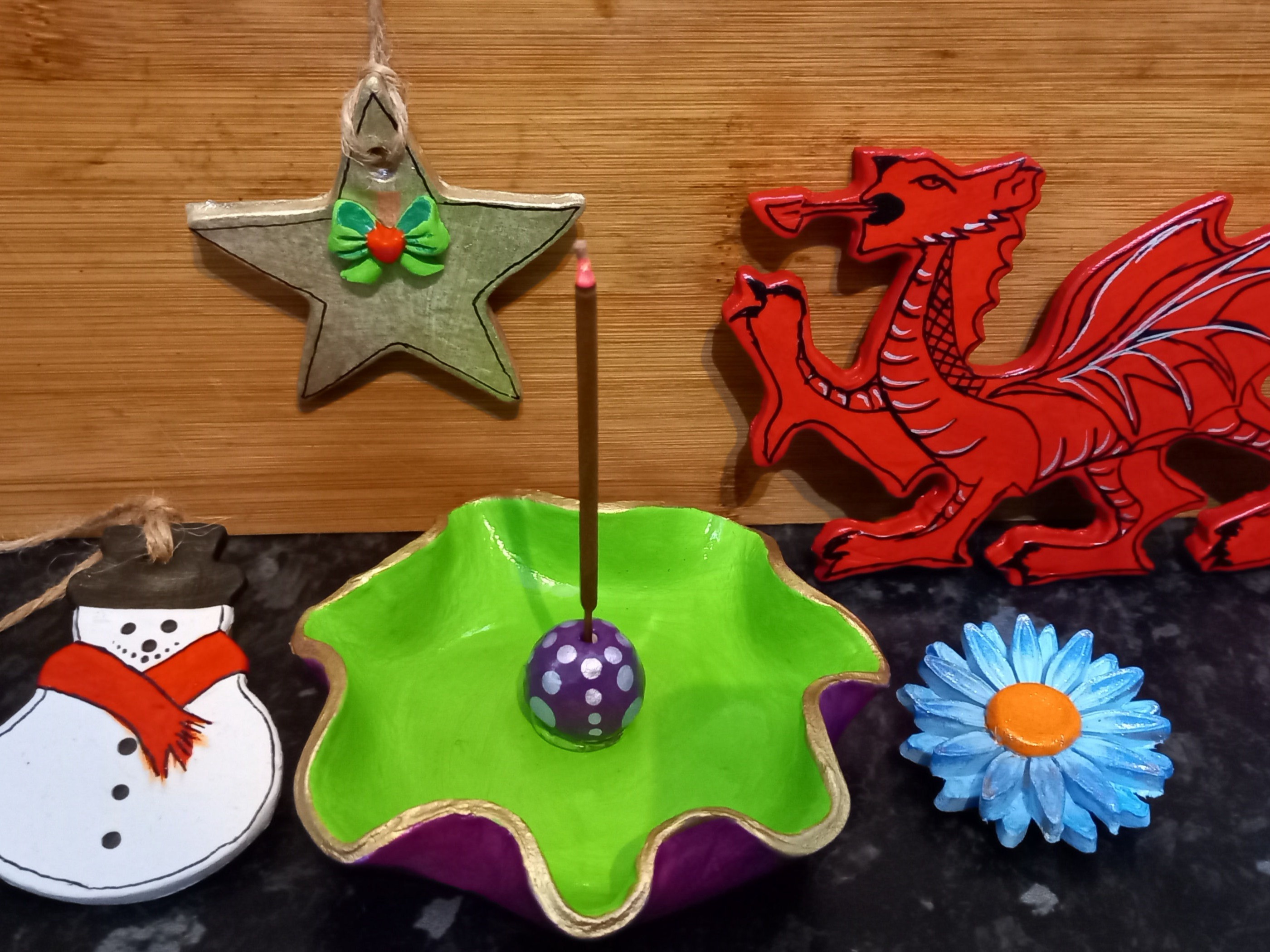 A colourful display features air dried clay pieces of the Welsh dragon, a green star, a purple and green incense holder, a snowman decoration, and a bright blue flower.