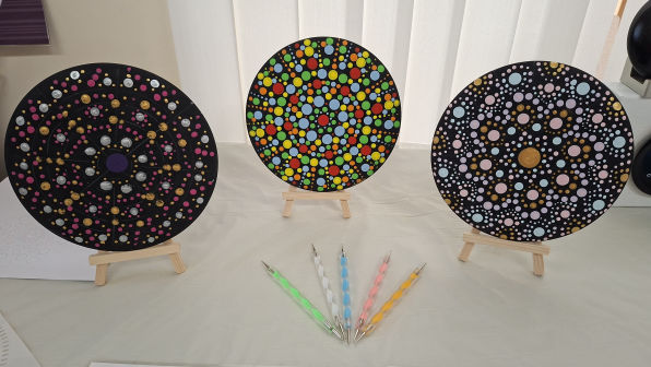 Three colourful mandala paintings on small easels display intricate dot patterns in vibrant colours. Below, six dotting tools are arranged in a fan shape.