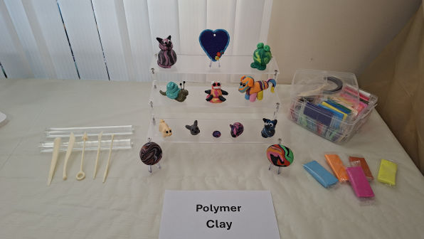 Display of colourful polymer clay figurines on a tiered stand, tools and clay blocks nearby. Label reads "Polymer Clay." Bright, creative atmosphere.