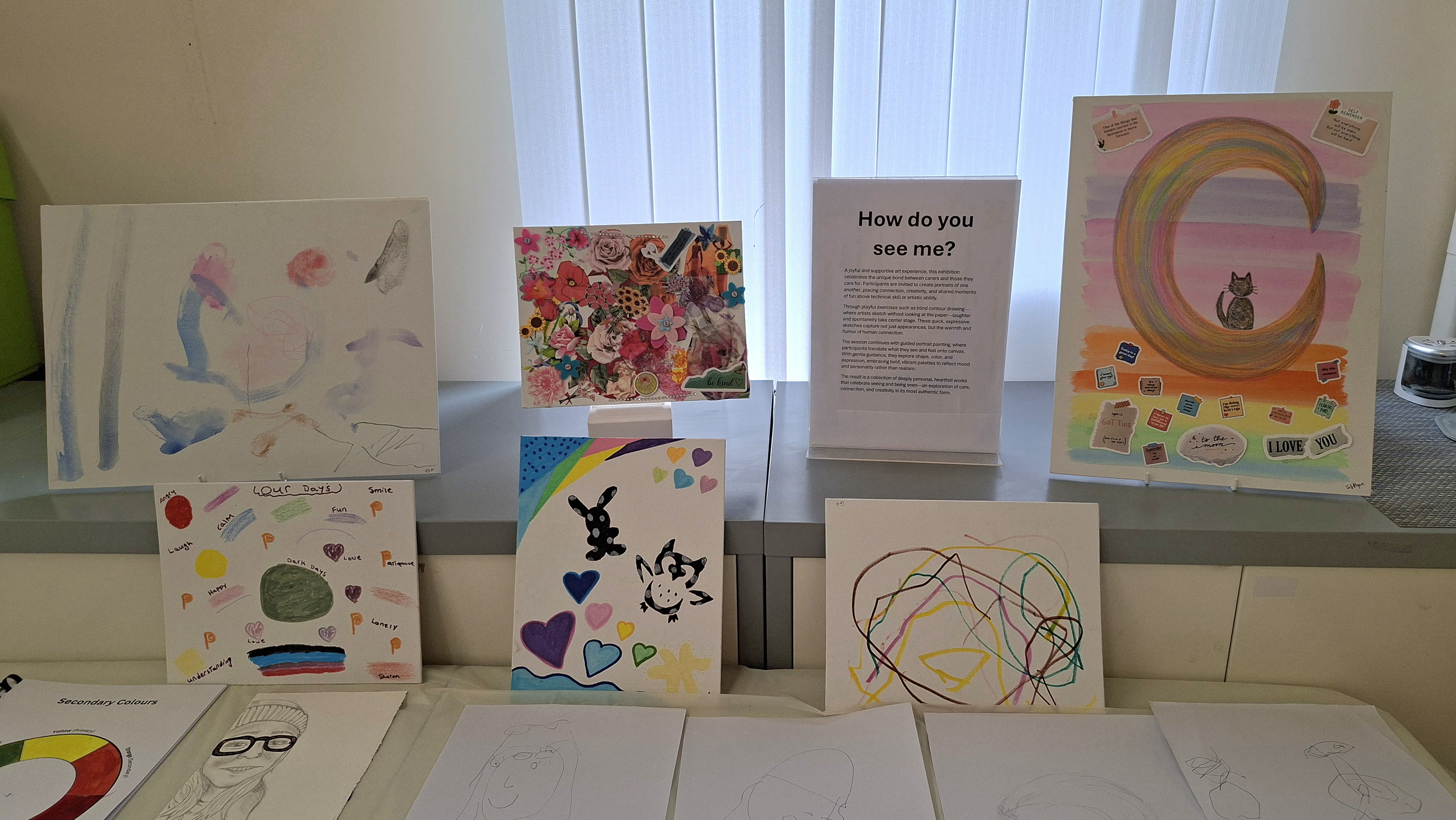 A collection of colorful artwork on display, featuring abstract designs, hearts, a cat on a crescent moon, and a floral collage. A sign asks, "How do you see me?" The artworks evoke creativity and introspection.