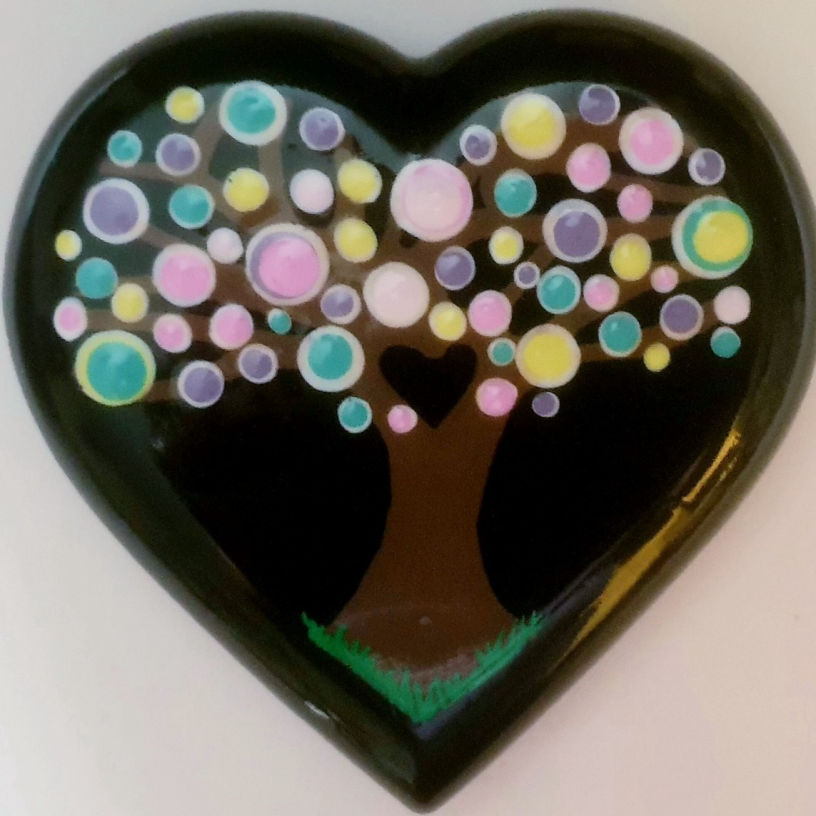 Heart-shaped design with a stylized tree of life. The tree has a brown trunk and colourful circular leaves in pink, yellow, and blue, on a black background.