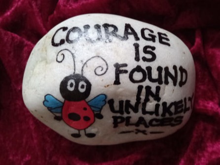 A painted rock on a red velvet background features a cute ladybug and the phrase, "Courage is found in unlikely places," evoking inspiration.