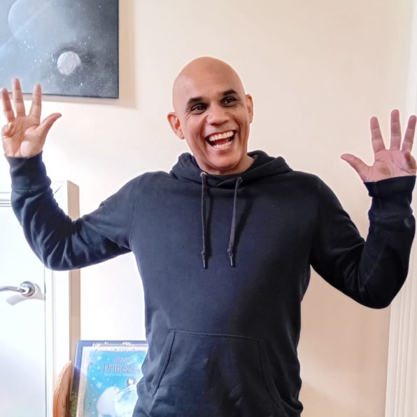 Darryl wearing a black hoodie standing in Smyrna with arms raised playfully. A framed space-themed picture and a book are visible in the background.