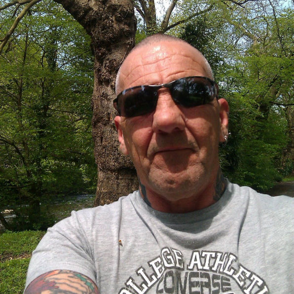 An image of Martin in sunglasses and a gray T-shirt poses outdoors with a calm expression. Background features lush green trees and a large tree trunk.