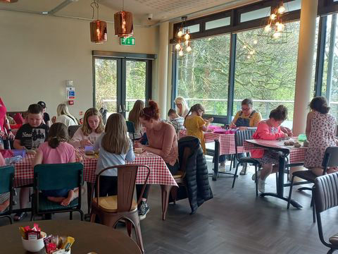 A lively café scene with people seated at tables with red chequered tablecloths, painting rocks. Large windows reveal a lush, green outside view.